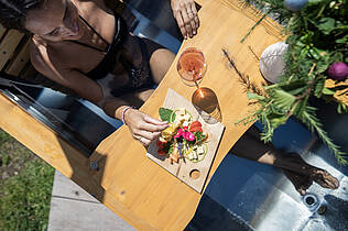 Frau liegt in der Outdoor-Badewanne und genießt würzigen Almkäse und ein Glas Frizzante beim AlmBad Erlebnis