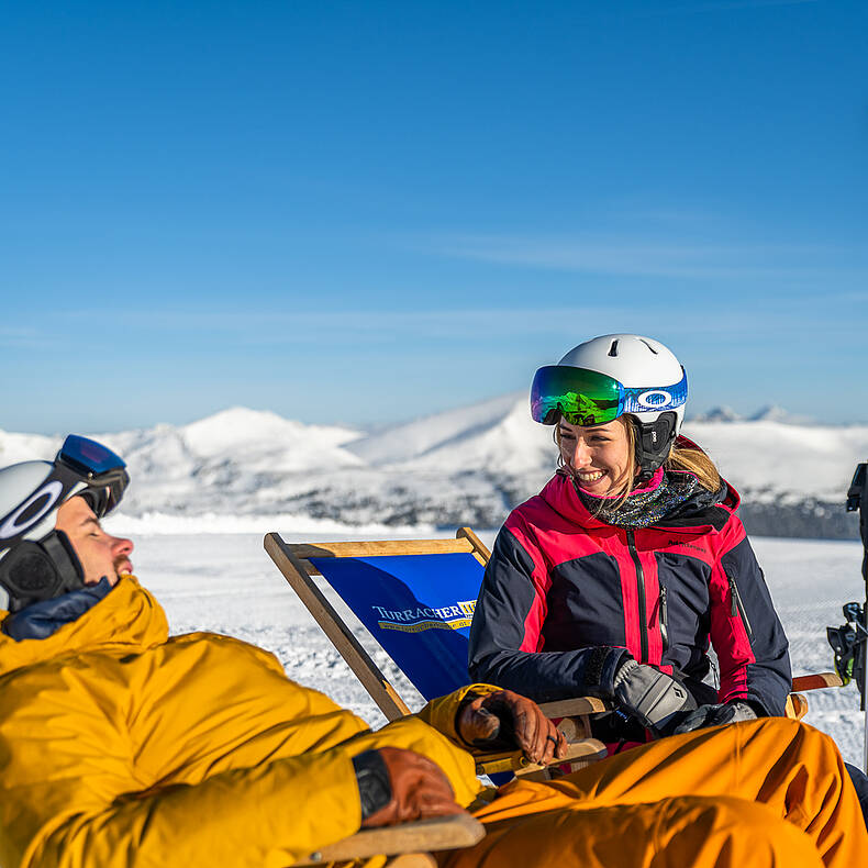 2 Personen sitzen in Liegestühlen, genießen die Sonne und haben Spaß miteinander