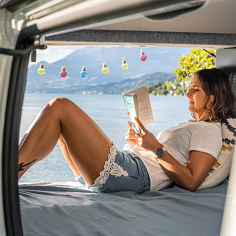 Frau ließt im Camper ein Buch mit See im Hintergrund