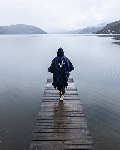 Person spaziert bei Regen mit Regenponcho bekleidet auf einem Steg Richtung See hinaus