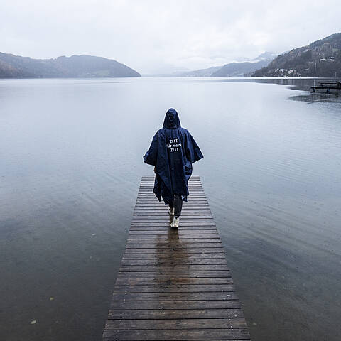 Person spaziert bei Regen mit Regenponcho bekleidet auf einem Steg Richtung See hinaus