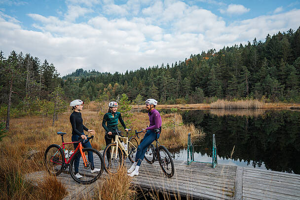 Drei Gravelbiker stehen mit ihren Fahrrädern auf einem Holzsteg am Egelsee in den Nockbergen und genießen die herbstliche Landschaft.
