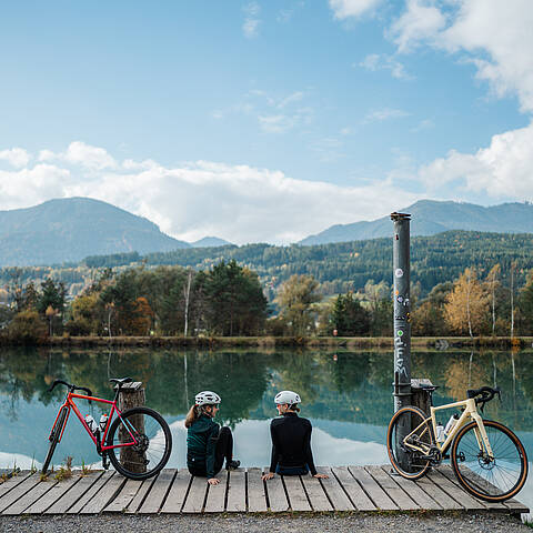 2 Personen rasten am Ufer der Drau mit ihren Gravel Bikes