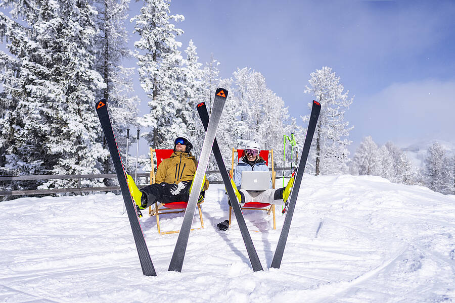 2 Personen sitzen mit einem Laptop in Liegestühlen und haben ihre Skier aufgestellt