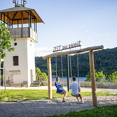 Ein Paar sitzt auf der Doppelschaukel „ZEIT zu zweit“ neben einem weißen Aussichtsturm im Barbara-Egger-Park. Dahinter liegen der Millstätter See und bewaldete Berge.