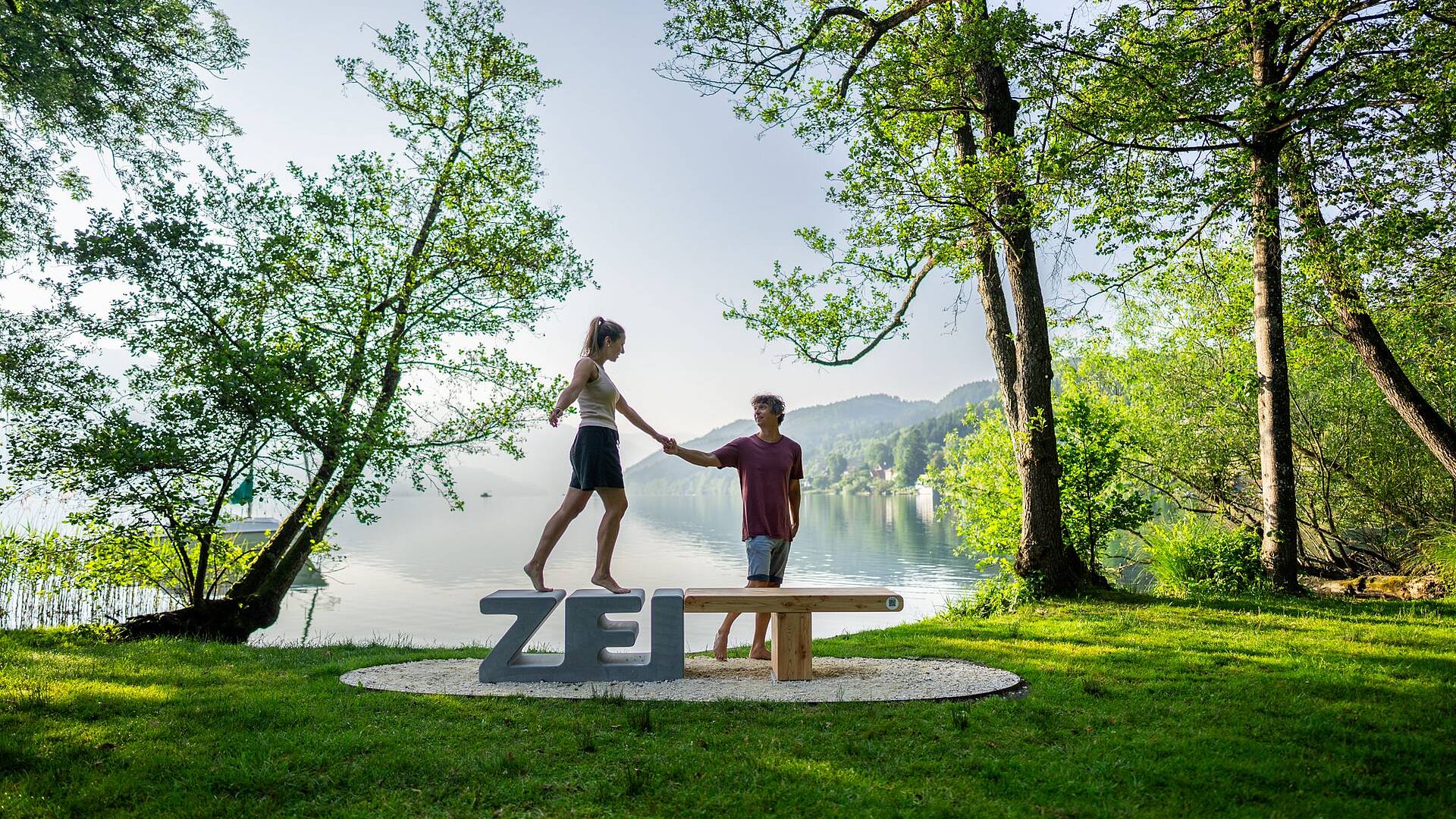 Frau balanciert auf Zeitbank in Seeboden und Mann steht daneben und hält sie