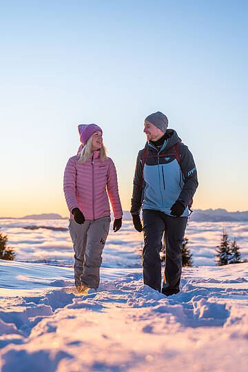 Zwei Personen gehen durch die verschneite Winterlandschaft der Nockberge bei tief stehender Sonne.