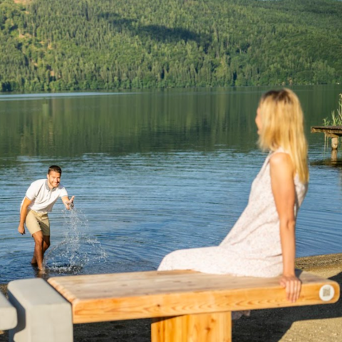 Frau sitzt auf ZEITbank, Mann steht im Millstätter See und spritzt mit Wasser