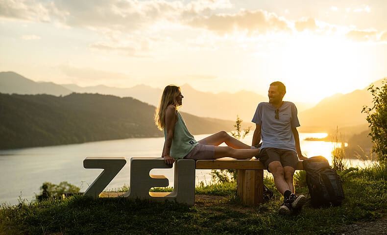 Paar sitzt auf der ZEITbank am ZEITplatz „Im Wandel der ZEIT“ mit Panoramablick auf den Millstätter See.