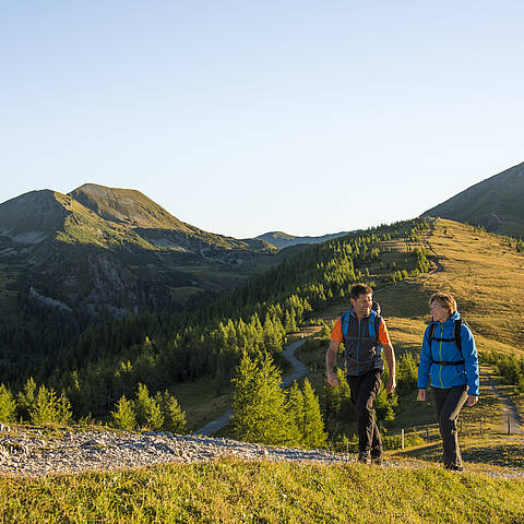  ALT-Text: Familie mit Kindern bei einer Wanderung in den Nockbergen, die glücklich an einem Gipfelkreuz steht und die Aussicht genießt. Bild 2: Wandern in den sanften Nockbergen Bildbeschreibung: Zwei Wanderer, ein Mann und eine Frau, gehen auf einem schmalen Pfad entlang eines grasbewachsenen Bergrückens in den Nockbergen. Der Mann trägt einen orangefarbenen Rucksack und eine graue Weste, die Frau eine blaue Jacke. Sie unterhalten sich und genießen die Bewegung in der Natur. Die Landschaft ist geprägt von sanften, grünen Hügeln und im Hintergrund sind weitere Gipfel der Nockberge zu sehen. Die Sonne scheint und taucht die Szene in ein warmes, goldenes Licht, das die friedliche und idyllische Atmosphäre des Wanderns unterstreicht. ALT-Text: Zwei Wanderer auf einem Wanderweg in den sanften, grasbewachsenen Nockbergen bei sonnigem Wetter.