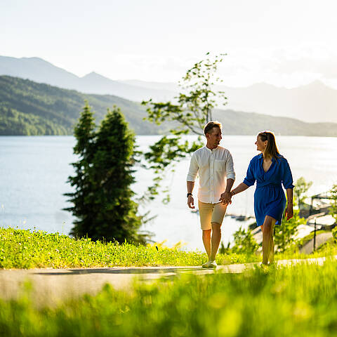 Ein Paar spaziert Hand in Hand auf einem sonnigen Weg oberhalb des Millstätter Sees, mit grünen Wiesen und Bergen im Hintergrund.