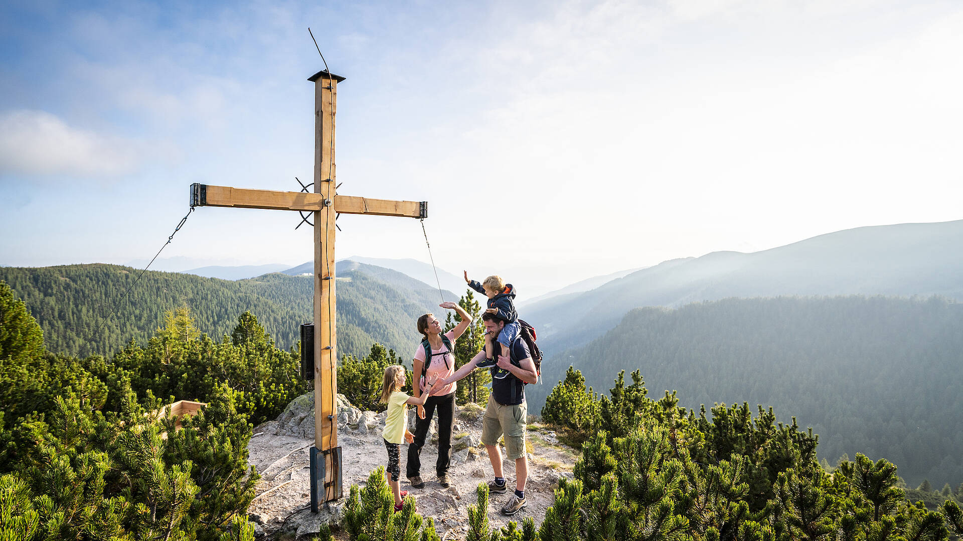Familie mit Kindern bei einer Wanderung in den Nockbergen, die glücklich an einem Gipfelkreuz steht und die Aussicht genießt.