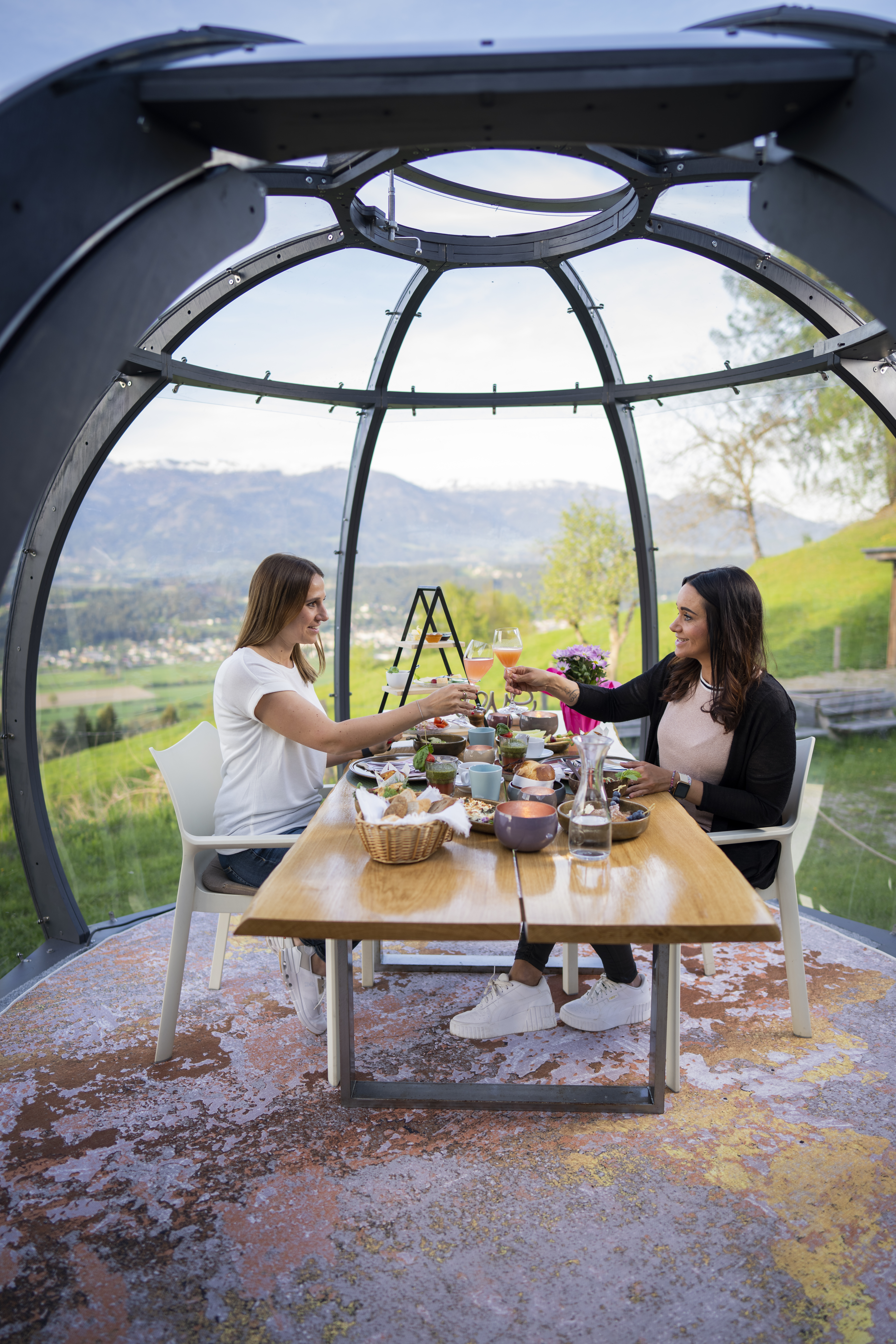 2 Frauen sitzen an einem gedecktem Tisch in der Glaskuppel und stoßen mit zwei Getränken an