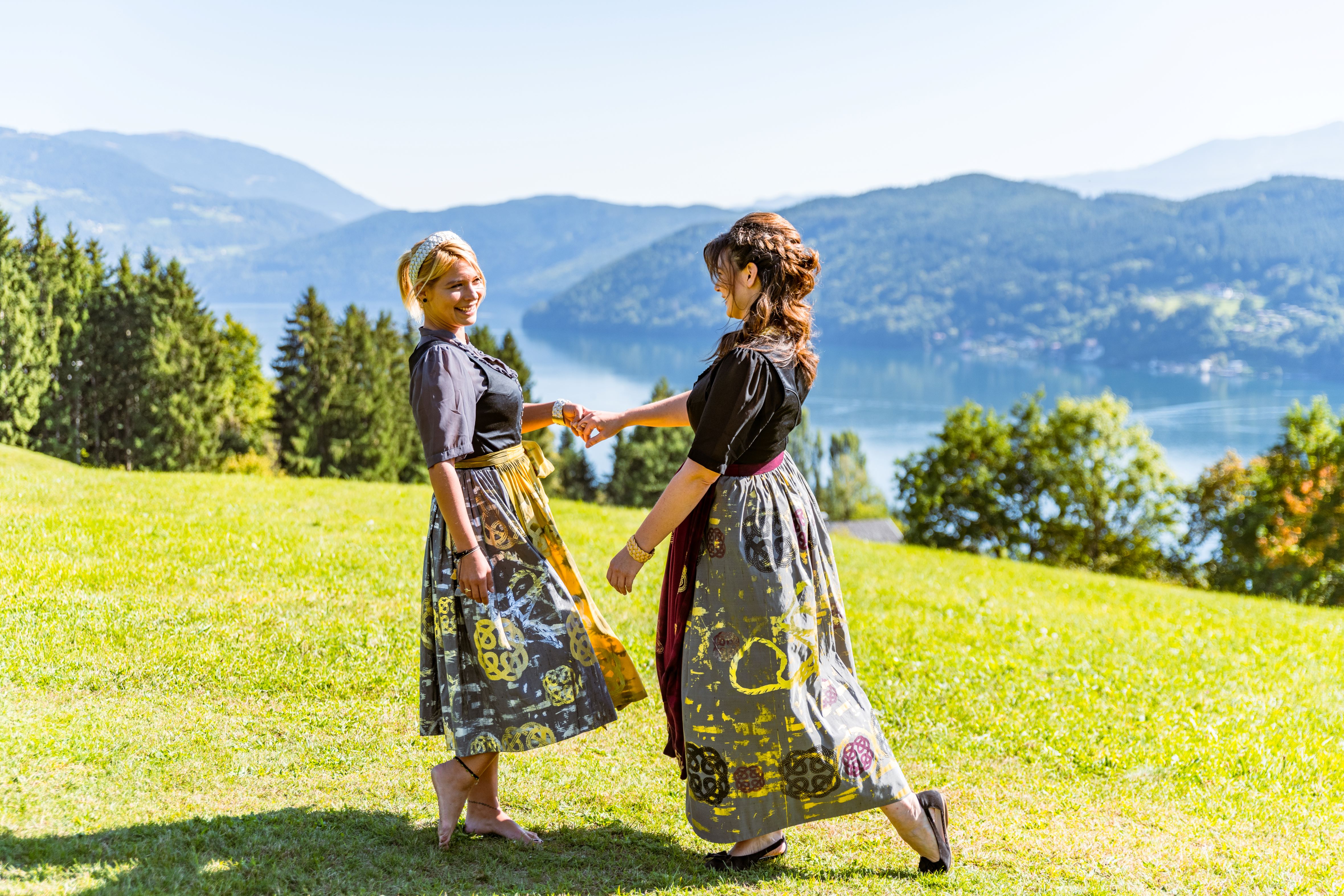 2 Frauen tanzen auf einer Wiese in Tracht mit Blick auf den Millstätter See